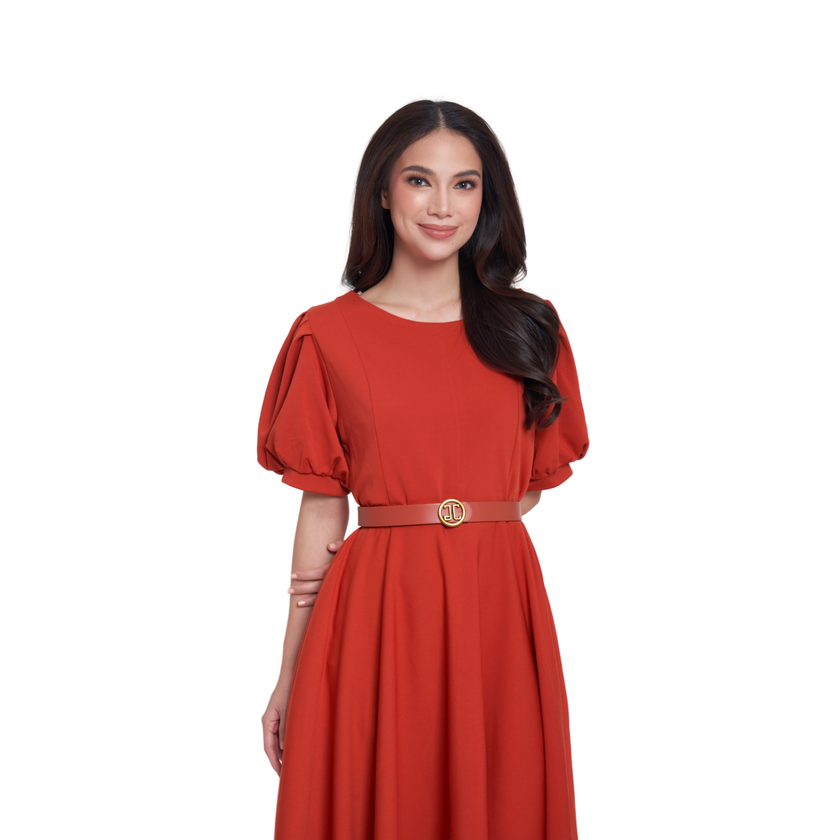 A woman modeling a rust-red midi dress with short puff sleeves, a round neckline, and a matching belt with a gold-tone circular buckle, set against a plain white background.
