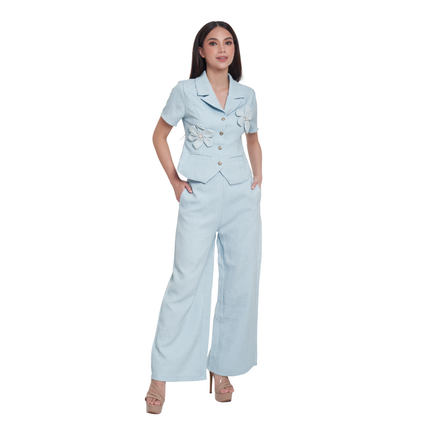A woman modeling a light blue two-piece suit consisting of a short-sleeve blazer with decorative fabric flowers and buttons, paired with matching wide-leg trousers, on a solid white background.