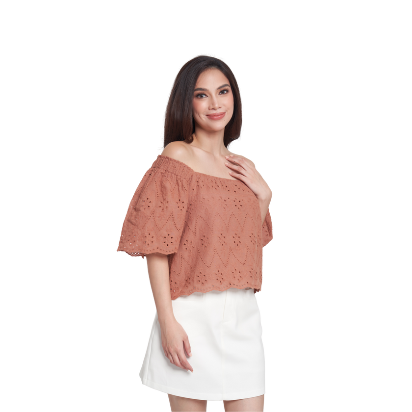 A woman modeling a terracotta-colored off-the-shoulder eyelet lace top with short flutter sleeves, paired with a white skirt, against a plain white background.