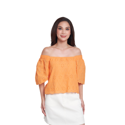 A woman modeling an off-the-shoulder eyelet top in a bright orange color, paired with a white skirt, against a plain white background.