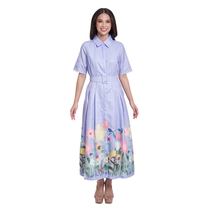 A woman modeling a light purple short-sleeve collared maxi dress with a matching belt and a colorful floral print along the hem, set against a plain white background.