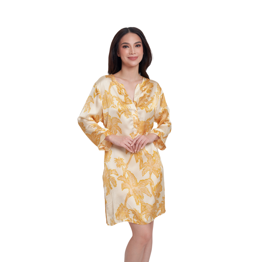 A woman modeling a short, V-neck satin dress with a yellow and gold palm leaf pattern on a cream background, set against a plain white studio backdrop.