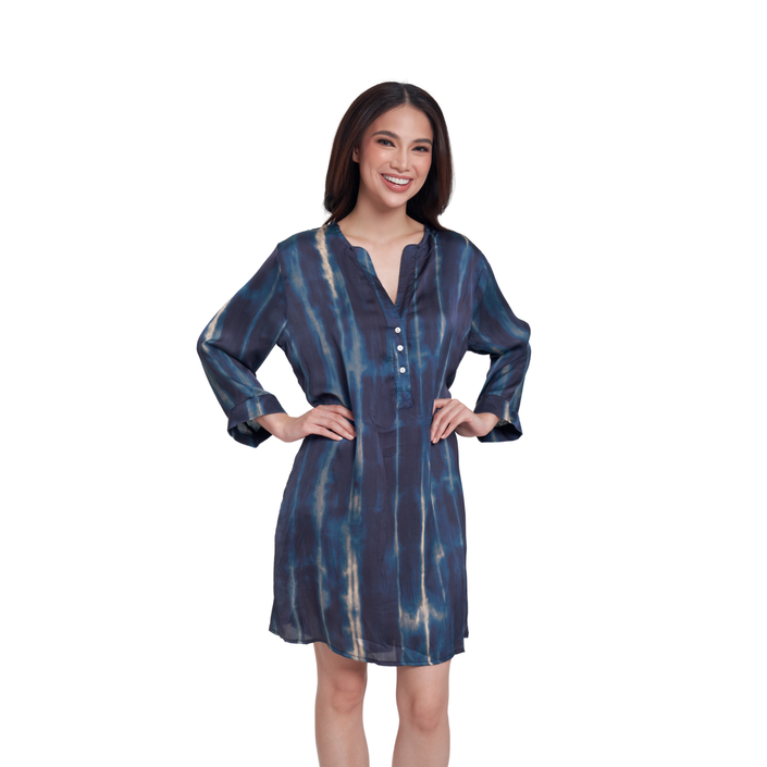 A woman modeling a short-sleeved, V-neck tunic dress with a blue and beige tie-dye pattern, standing against a solid white background.