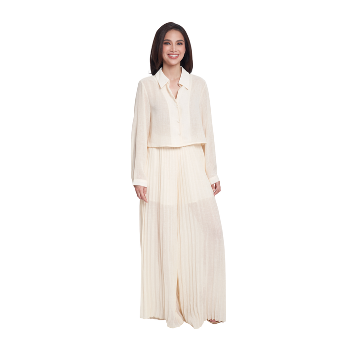 A woman modeling a cream-colored two-piece outfit consisting of a long-sleeve collared blouse and a matching pleated maxi skirt, set against a plain white background.