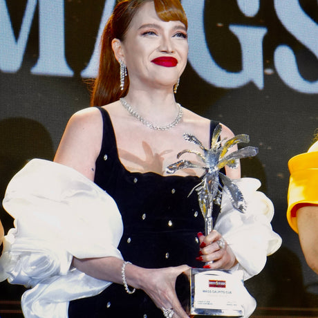 A woman on stage holding a silver award trophy, wearing a black velvet dress with white puffed sleeves and silver embellishments. The background features a dark screen with large light blue letters.