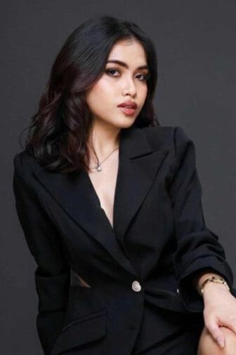 A woman wearing a black blazer and matching shorts sits on a white block against a dark gray studio background.