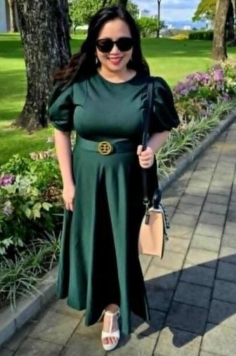 A woman wearing a dark green midi dress with puff sleeves and a gold belt, standing on a paved path in a park with trees and green grass in the background.