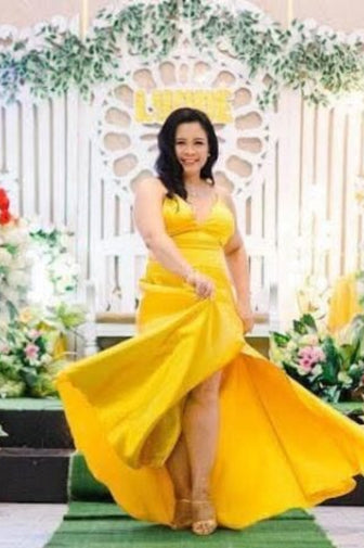 A woman wearing a bright yellow formal gown with a high-low hemline walks down a green carpet in front of a decorated white arch with floral arrangements.