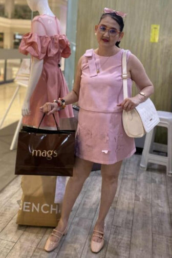 A woman wearing a light pink sleeveless mini dress and matching pink flats stands in a boutique holding a brown French Bulldog shopping bag and a white shoulder bag.